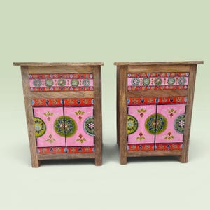 Op de afbeelding: Twee houten nachtkastjes met een frame van natuurlijk hout, een lade en twee kastdeurtjes. De voorpanelen zijn roze geschilderd met groene, rode en witte cirkelvormige bloemmotieven en decoratieve randen op de lade en de deurtjes.