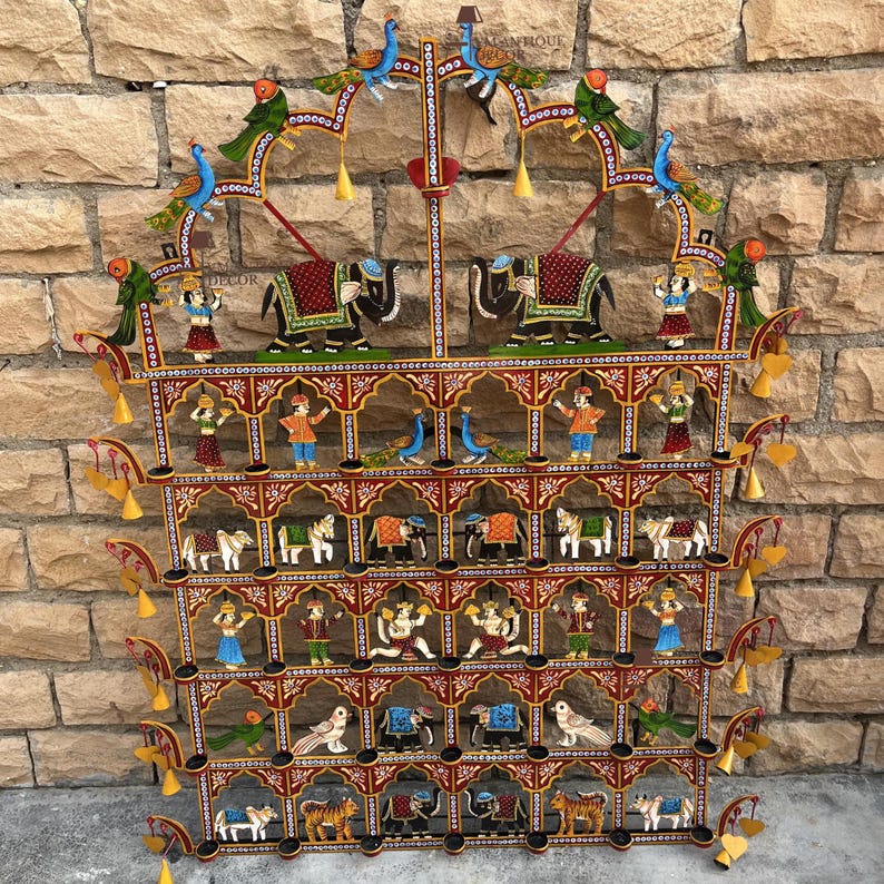 Puede incluir: Un colgante de pared de metal de varios niveles con detalles pintados a mano de elefantes, tigres, aves y figuras humanas. La estructura incluye compartimentos arqueados, adornos de campanas colgantes y colores vibrantes en rojo, oro y azul.