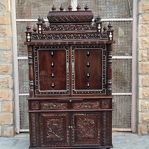 K&ouml;nnte beinhalten: Ein dunkelbrauner Holzschrank mit aufwendigen Schnitzereien und einer detaillierten Oberseite. Der Schrank hat zwei T&uuml;ren, dekorative Akzente und kleine Schubladen. Der Schrank steht auf Rollen und hat ein traditionelles Design.