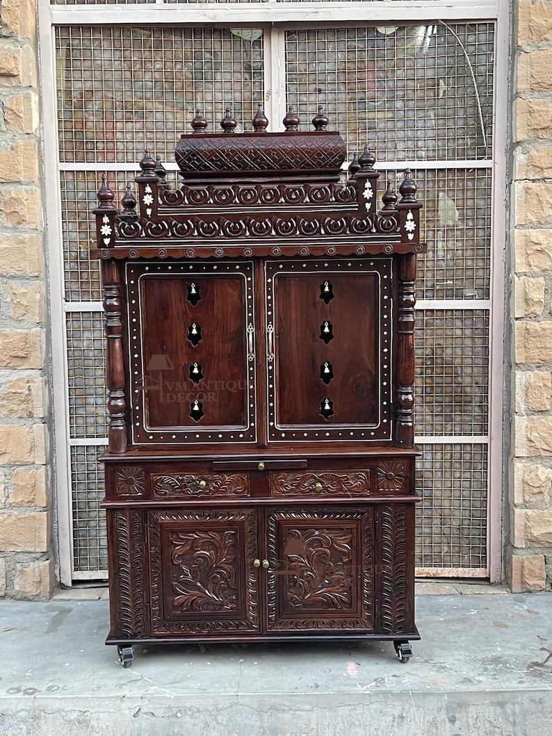 K&ouml;nnte beinhalten: Ein dunkelbrauner Holzschrank mit aufwendigen Schnitzereien und einer detaillierten Oberseite. Der Schrank hat zwei T&uuml;ren, dekorative Akzente und kleine Schubladen. Der Schrank steht auf Rollen und hat ein traditionelles Design.