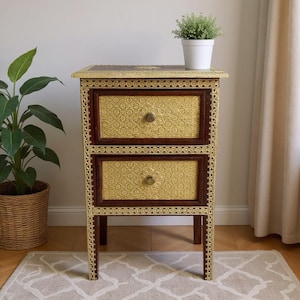 May include: A rectangular wooden bedside table featuring two drawers with embossed gold-colored metal panels. The frame is accented with intricate gold-colored metal trim along the edges and legs. Each drawer has a single round gold-colored knob.