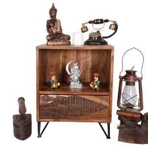 May include: A wooden nightstand with a single drawer and black metal legs, topped with a bronze Buddha statue, a white candle, and a black rotary telephone. Inside the shelf are two small colorful figurines and a silver crescent moon sculpture.