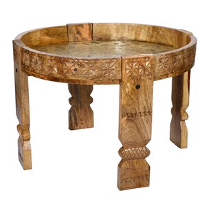 May include: A round wooden side table featuring a natural light brown finish with carved geometric patterns along the outer rim. The tabletop surface is inlaid with a circular brass plate displaying intricate etched designs, supported by four legs.