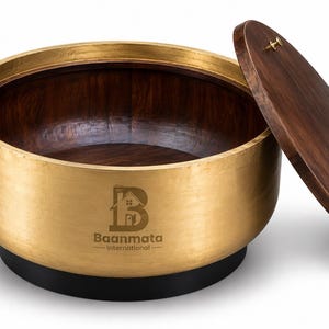 Pode incluir: Um recipiente redondo com exterior dourado metálico, interior em madeira castanho-escura e uma tampa de madeira castanho-escura a condizer. O lado apresenta o logótipo e o texto Baanmata International. A base é preta e a tampa tem um pequeno puxador de latão.