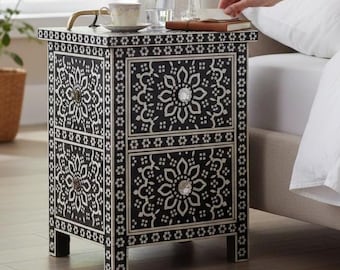 May include: A rectangular bedside table featuring a black finish with intricate white floral inlay patterns. The piece includes two drawers, each equipped with a clear, faceted crystal-style knob, and stands on four short, straight legs.