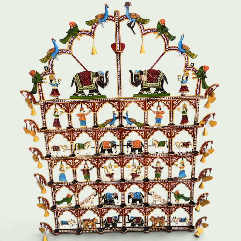 Puede incluir: Un adorno de pared de metal de varios niveles con detalles pintados a mano de elefantes, tigres, vacas, pavos reales y figuras humanas. La estructura est&aacute; decorada con peque&ntilde;as campanas colgantes y arcos ornamentados en tonos rojo, azul y oro.