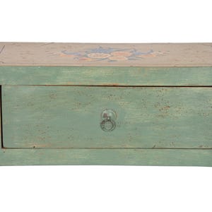 May include: A rectangular wooden chest with a distressed sage green finish, featuring a single central drawer with a circular metal pull. The top surface is painted with a floral motif in muted tones, and black metal handles are on the sides.