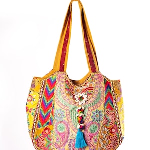 Könnte beinhalten: Eine gelbe Tragetasche mit aufwendigen, mehrfarbigen Stickereien, Patchwork-Mustern und Pailletten-Akzenten. Die Tasche verfügt über ein zentrales Dekorelement aus weißen Kaurimuscheln und eine leuchtend blaue Quaste an der Vorderseite.