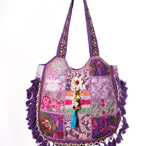 Könnte beinhalten: Eine violette Patchwork-Tragetasche mit verschiedenen floralen und geometrischen Stickmustern. Die Tasche ist mit violetten Quasten an den Seiten und am Boden sowie einem zentralen Anhänger aus weißen Kaurimuscheln und bunten Perlen verziert.