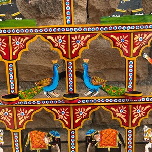 Puede incluir: Un colgante de pared de metal de varios niveles con elefantes negros pintados a mano con mantas naranjas, pavos reales azules con colas verdes y motivos florales amarillos. La estructura incluye varios portavelas circulares peque&ntilde;os.