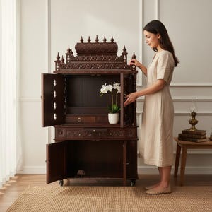 K&ouml;nnte beinhalten: Ein dunkelbrauner Holzschrank mit kunstvollen Schnitzereien, offenen T&uuml;ren und einer kleinen Schublade. Der Schrank hat eine dekorative Oberseite und steht auf Rollen. Eine wei&szlig;e Orchidee im Topf befindet sich im Inneren.