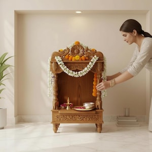 May include: A carved wooden home temple featuring intricate floral and peacock motifs, adorned with a white jasmine garland and orange marigold flowers. The structure includes a central shelf holding a small brass lamp and a silver bowl.