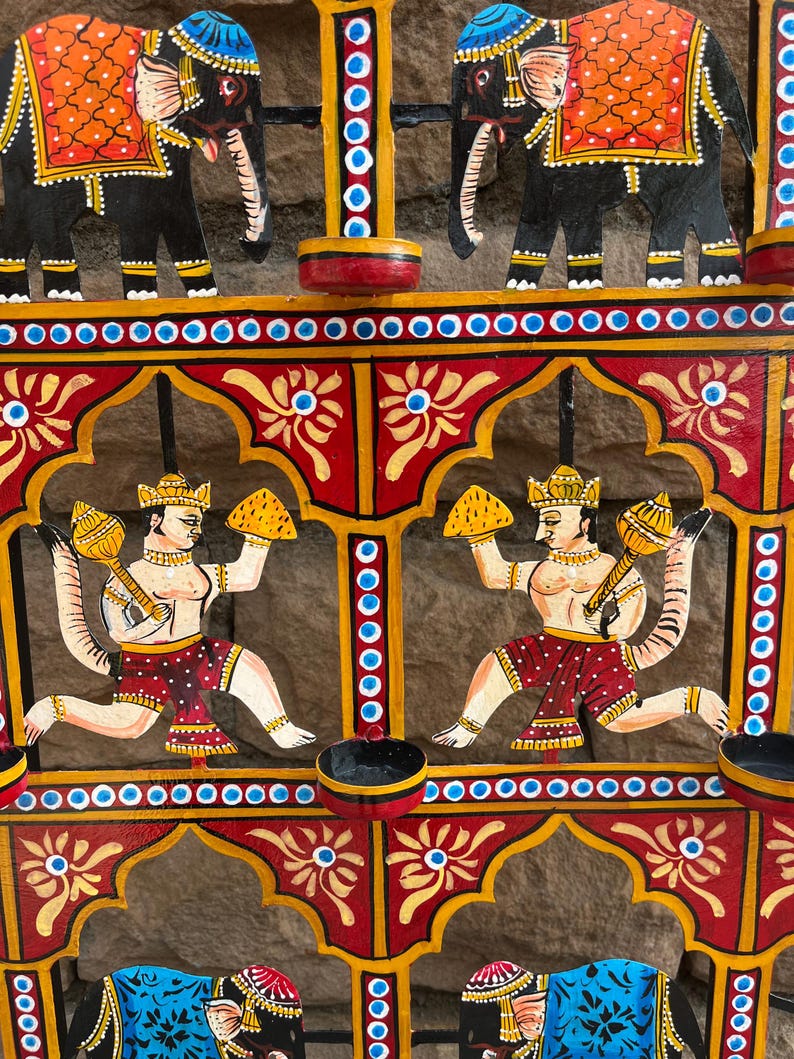 Puede incluir: Un colgante de pared decorativo de metal con elefantes negros pintados a mano con mantas naranjas y dos figuras con colas que sostienen mazas doradas. El marco est&aacute; pintado en rojo, amarillo y azul con motivos florales y portavelas.