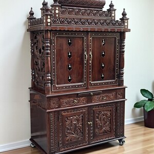 K&ouml;nnte beinhalten: Ein dunkelbrauner Holzschrank mit aufwendigen Schnitzereien und Intarsien. Der Schrank hat mehrere T&uuml;ren, Schubladen und dekorative Zierelemente. Er steht auf Rollen und hat ein traditionelles Design.
