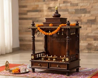 May include: A dark brown wooden home temple featuring four carved pillars, a tiered roof with a central finial, and two storage drawers. The structure is decorated with an orange marigold garland and small brass bells, with lit oil lamps inside.