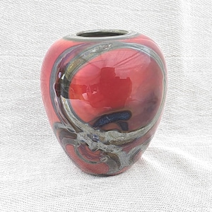 May include: A rounded glass vase featuring a glossy red base colour with swirling, abstract patterns in shades of dark blue, grey, and metallic silver. The vessel has a smooth, reflective surface and a circular opening at the top with a dark rim.