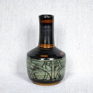 May include: A ceramic vase with a dark brown and black glazed finish. The vase has a cylindrical neck and a rounded body with a green and black pattern. The base is a lighter brown color.
