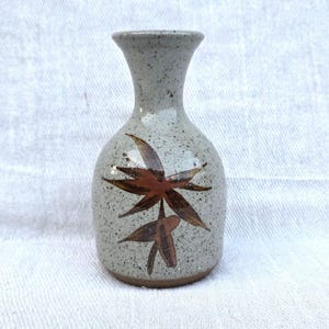 May include: A small, speckled ceramic vase with a narrow neck and rounded body. The vase features a brown leaf design. The vase is approximately 13 cm tall.