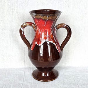 May include: A vintage ceramic vase with two handles. The vase has a glossy brown base, transitioning to a vibrant red and white centre, and a brown rim. The design is reminiscent of lava flow. A decorative piece for home decor.