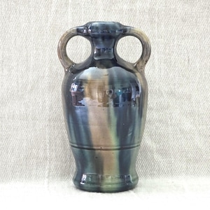 Peut inclure: Un vase en céramique avec deux anses en boucle. Le vase a une finition émaillée bleu foncé, marron et verte. Le vase a un corps arrondi et une base évasée. Les anses sont circulaires et fixées au sommet du vase.