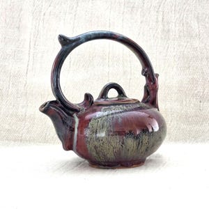 Può includere: Una piccola teiera in ceramica fatta a mano con una glassa rosso scuro, verde e nera. La teiera presenta un corpo arrotondato, un piccolo beccuccio e un manico grande e arcuato. Il coperchio ha un piccolo pomello. Il design è rustico e artistico.