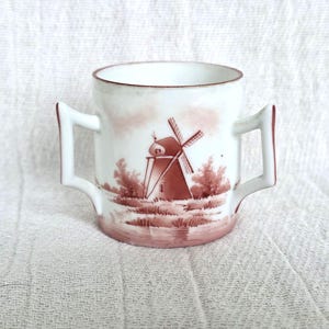 Puede incluir: Taza de cerámica blanca con dos asas, con una ilustración en tonos sepia de un molino de viento y un paisaje. El borde y los bordes de las asas son de color marrón oscuro. La taza está sobre una superficie blanca texturizada.