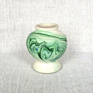 May include: A small, bulbous ceramic vase featuring a marbled pattern of various shades of green, including forest, lime, and mint, with subtle streaks of blue. The vase has a smooth, rounded body, a narrow neck, and a flared base in a matte off-white.
