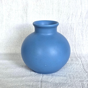 Könnte beinhalten: Eine kleine, runde, blaue Vase mit schmalem Hals. Die Vase hat eine matte Oberfläche und steht auf einer weißen, strukturierten Oberfläche. Diese dekorative Vase eignet sich zum Präsentieren von Blumen oder als eigenständiges Akzentstück.