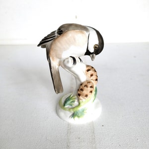 Puede incluir: Una figurita de porcelana de un pájaro pequeño posado en una rama con hojas verdes. El pájaro es marrón, blanco y negro con un ojo negro. La figurita está sobre un fondo blanco.