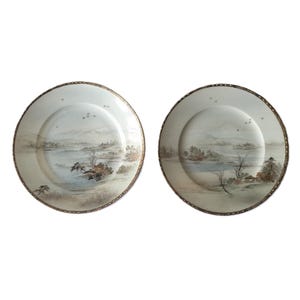 Può includere: Due piatti in ceramica bianca con bordo dorato. Ogni piatto presenta una scena di paesaggio dipinta a mano con montagne, alberi e un lago. La scena è dipinta in tonalità di marrone, grigio e blu.