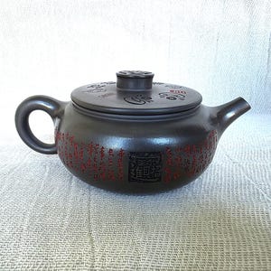 Pode incluir: Bule de cerâmica marrom escuro com tampa a condizer. O bule apresenta um corpo arredondado, uma alça curva e um bico. Caracteres chineses vermelhos estão inscritos ao redor do corpo. A tampa tem um pequeno botão e gravuras decorativas.