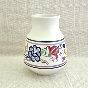 Könnte beinhalten: Weiße Keramikvase mit rundem Körper und ausgestelltem Rand. Die Vase hat ein dekoratives Band mit floralen Designs in Blau, Lila und Rot, akzentuiert mit grünen und gelben Details. Zwei grüne und violette Streifen umrunden die Vase.