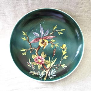 May include: A round ceramic plate with a teal green glaze and a white rim. The plate is decorated with a floral design featuring pink, yellow, and purple flowers, green leaves, and brown branches. The plate is a decorative item.