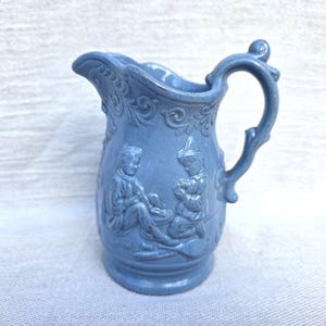 May include: A light blue ceramic jug with a detailed handle and spout. The jug features relief carvings of figures in a pastoral scene. The jug is approximately 15 cm tall.
