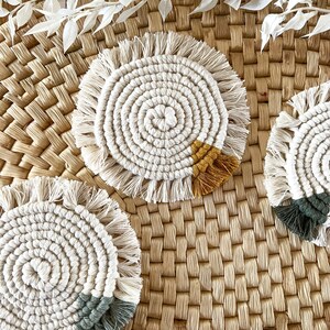 May include: Three round crocheted coasters with fringe. The coasters are white with a single colored accent at the bottom. One coaster has a yellow accent, one has a green accent, and one has a blue accent. The coasters are sitting on a woven straw surface.