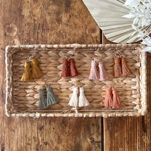 May include: Six pairs of tassel earrings in a woven basket. The earrings are in various colors, including mustard yellow, burnt orange, light pink, sage green, cream, and terracotta.