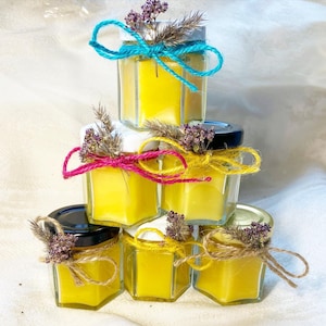 Pode incluir: Uma pilha de seis potes hexagonais de vidro cheios de velas de cera amarela. Cada pote é adornado com um laço de barbante colorido e flores secas. Os potes têm tampas pretas, brancas e douradas.
