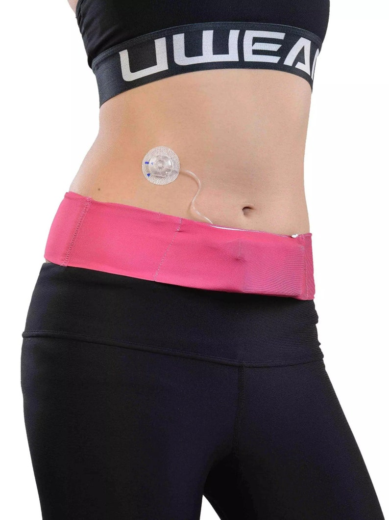 May include: A close-up of a person's midsection, showcasing a pink waist belt and black leggings. A medical device is attached to the abdomen. The person is wearing a black sports bra with the word "UWEAR" printed on it.