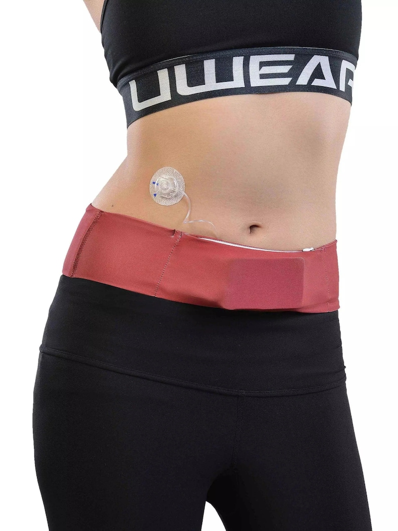 May include: A person wearing a black sports bra and black leggings. A red-brown waist belt with a zipper is worn around the waist. A medical device is attached to the abdomen.