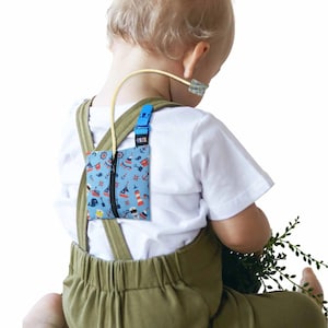 Könnte beinhalten: Eine blaue Tasche mit nautischem Muster, mit Ankern, Segelbooten und Leuchttürmen, ist mit einem blauen Clip an den Hosenträgern eines Kindes befestigt. Die Tasche hat einen schwarzen Reißverschluss und ist mit einer Ernährungssonde verbunden.