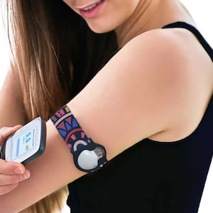 Puede incluir: Una mujer lleva un sujetador deportivo negro con una banda para el brazo de estampado colorido y un pequeño dispositivo blanco unido a él. Está sosteniendo un pequeño dispositivo negro con una pantalla que muestra "5.5".