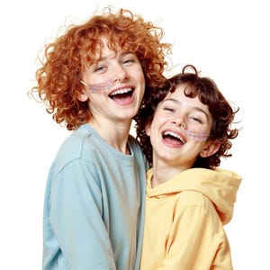 Op de afbeelding: Twee mensen die lachen en lachen, gekleed in lichtblauwe en gele sweatshirts. Elke persoon heeft een kleurrijk patroonverband op hun gezicht. De achtergrond is wit.