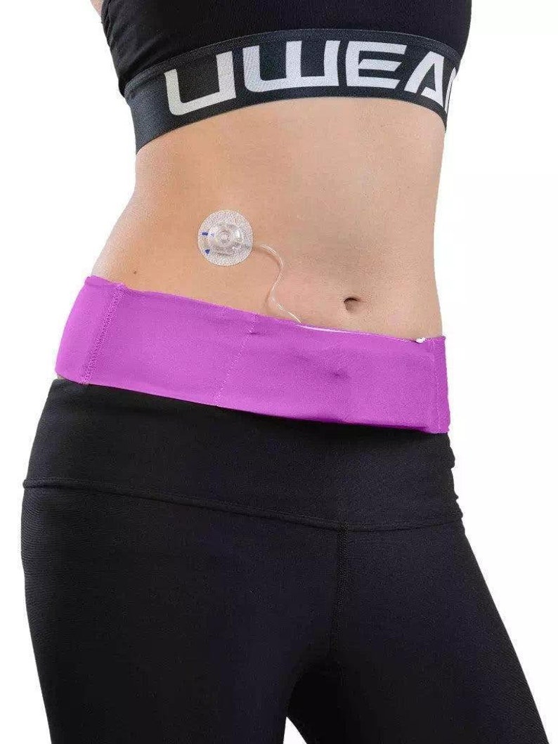 May include: A person wearing a black sports bra with the word "UWEA" printed on it, black leggings, and a purple waist belt. A medical device is attached to the abdomen.