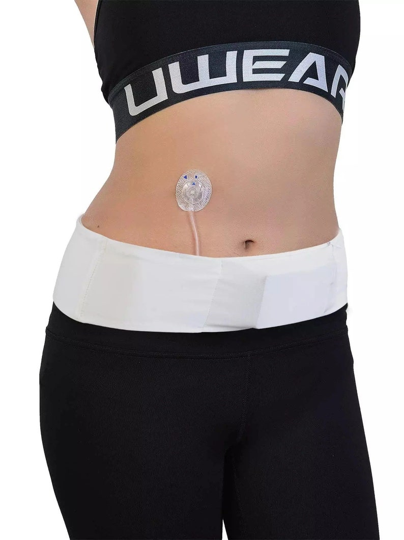 May include: A white medical belt worn around the waist, paired with black leggings and a black sports bra. A clear medical device is attached to the abdomen. The sports bra has the word "UWEAR" printed on it.