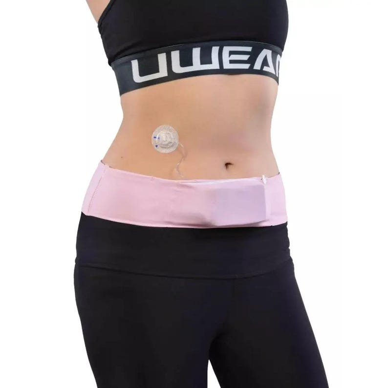 May include: A pink waist belt with a small pocket, worn over black athletic leggings. A black sports bra with white text is visible above the belt. A medical device is attached to the abdomen.