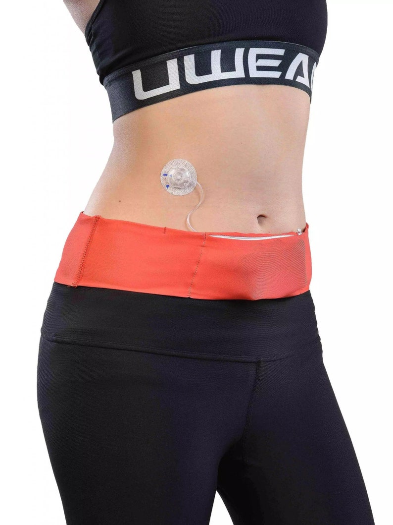 May include: A person wearing a black sports bra and black leggings. An orange waist pack with a zipper is worn around the waist. A medical device is attached to the abdomen.