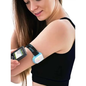 Può includere: Una donna indossa una canotta nera con un tracker fitness blu e nero sul braccio superiore. Sta tenendo un piccolo dispositivo nero con uno schermo che mostra del testo.