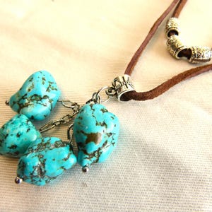May include: A turquoise stone necklace with a brown suede cord. The necklace features four turquoise stones with dark brown veining, connected by a silver chain. The cord has silver decorative beads.