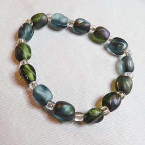 May include: A beaded bracelet featuring oval-shaped beads in shades of green, blue, and hints of purple. The beads are strung together with small, clear beads, creating a flexible and stylish accessory. The bracelet is displayed on a white surface.