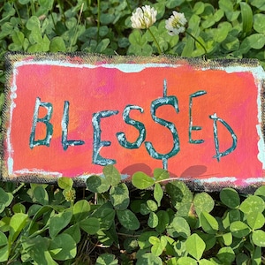 May include: A small, rectangular sign with a bright orange background and a black border. The word "BLESSED" is written in green paint on the sign.